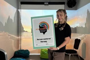 A young woman is standing in a room in front of a landscape-looking background. She is holding a banner with the organisation's logo and the contact details. In the middle of the banner it says 'Person-centered therapy' in big bold letters. She is wearing a T-shirt with the same logo printed on it.