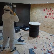 A person in a white protective suit and helmet swings a hammer inside a rage room, surrounded by smashed debris on the floor, with a stack of tyres and the words "Release the rage" painted in red on a wooden board wall behind them.