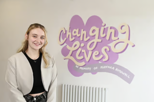 A blond young woman stands in front of a wall. The wall reads the organisations slogan: 'Changing Lives - In Memory of Aleysha Rothwell'