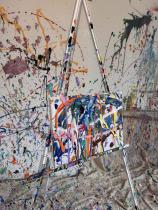 The picture shows a room with the walls and floor covered in white fabric. These are full of paint of different colours, like an artwork. In the centre of the room, an easel holds a canvas with paint strokes that look exactly like the walls. 