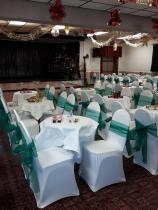 The venue shows several rows of empty tables and chairs. Christmas decorations hang from the ceiling and Christmas crackers lay on the tables. In the background there is a small stage.