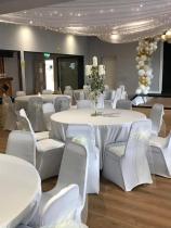 Four big round tables lay in a venue, surrounded by several chairs with white covers. There are candles on the table, dimmed fairy lights hanging from the ceiling and bunches of balloons in the background. The room looks prepared to host an event, such an anniversary or reception. 