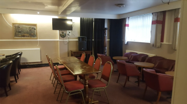 The image shows a room with three sections, all of them are laid out with a few low tables and numerous chairs around them. On the right hand side there are two windows, in the front wall there is a radiator, a painting, a TV and a speaker sitting on top of a stand.