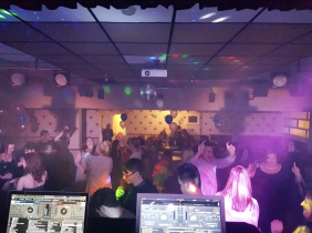 The image shows a venue with a big group of people dancing. There are balloons around. In the ceiling, a disco ball and a projector. The spotlights light the room. Two laptops display DJ software.