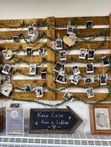 A wooden pallet-style structure is on the wall, displaying several pictures of the staff and customers at the cafe. On the below table there is a sign that says 'Keep Calm. Have A Coffee" and a small holder explaining the cafe's mission.
