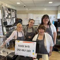 A group of four women of different ages stand behind a counter showing a cash register and, in the background, a kitchen. They smile to the camera and wear aprons with the cafe's logo.
