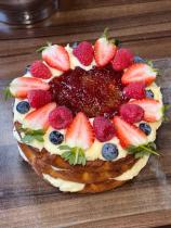 A close-up image of a rounded cake showing various layers of sponge and cream, has been decorated with strawberries, raspberries, blueberries and jam on top.