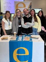 Four women and a man stand behind a table. They are smiling. The table displays the organisations leafleats during an event.