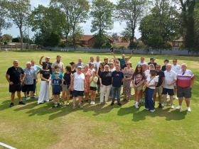 A group of 30 children and adults of different ages stand outdoors in a bowling field, cheering and posing for the picture.