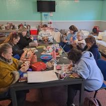 A group of eight middle-aged women are sat around a big table in a room that resembles a workshop. The group are making handmade items using the yarn, pegs and colour pencils that lay on the table.