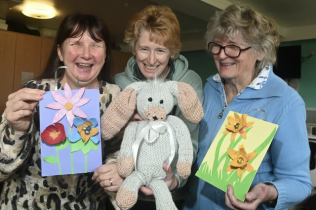 Portrair of three middle-aged women who smile and exhibit handmade items. Two of them are paper flowers and another one is a teddy bear.