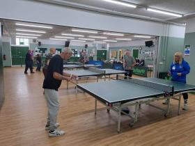 A group of ten middle-aged mixed-gender individuals play ping-pong in five ping-pong tables laid across a big indoors room.