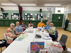 A group of eight middle-aged women smile and sit around a big table, drinking a hot drink and knitting.