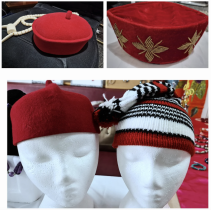 A collection of traditional cultural headwear displayed on mannequin heads, including a red embroidered cap, a plain red cap, and a black, white, and red knitted hat. The handcrafted designs showcase African heritage, cultural fashion, and artisanal craftsmanship.