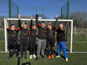 A group of 6 young people stand in a football pitch in front of the goal. It is a sunny day. The group hold their arms up and cheer. 