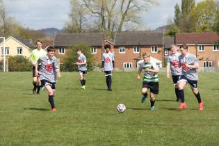 A group of teenage boys play football outdoors in a football pitch on a sunny say. Some of them are wearing the Wigan Cosmos football kit. A few houses and trees appear in the background.