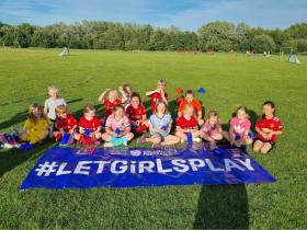 A group of 15 young girls dressed in sport wear are sat on the grass on a sunny day. In front of them lays a wide banner that reads: "England Foodball #LetGirlsPlay".