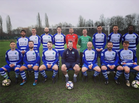 A group of eight men pose for their football group portrait outdoors, in a football pitch. They are wearing the Wigan Cosmos football kit.